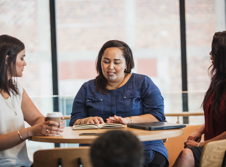 A group of three Māori women sit at a desk talking