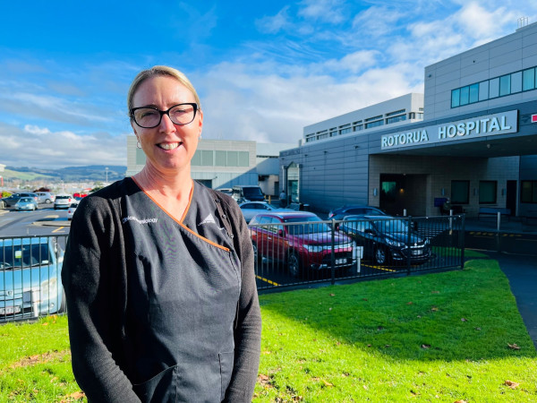 A white woman in black clinical physiology uniform stands outside the Rotorua Hospital main entrance. she is wearing glasses and smiling.