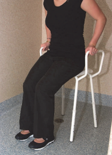 A woman sits of a white stool with arm grips inside a shower cubicle. The woman is lowering herself to sitting position as she grips the handles.s. 